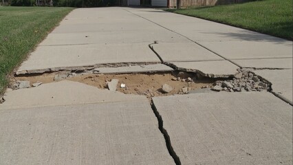 A crack in the sidewalk is shown with a few pieces of concrete and a few pieces of trash