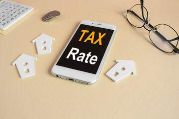 Fototapeta premium The expression Tax rate on the mobile phone screen. Beautiful beige background. Glasses, miniature houses, coins. Business concept.