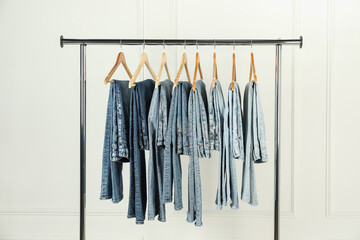 Many stylish jeans hanging from rack near white wall