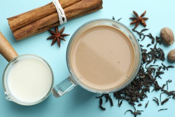 Aromatic Masala tea in glass cup, spices, dry leaves and milk on light blue background, flat lay