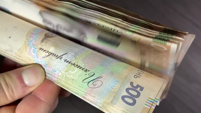 Hands are counting, throwing, and collecting a large stack of 500 hryvnia banknotes, with cash scattering on a laptop background.