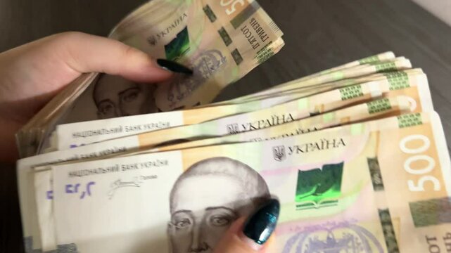 Hands are counting, throwing, and collecting a large stack of 500 hryvnia banknotes, with cash scattering on a laptop background.