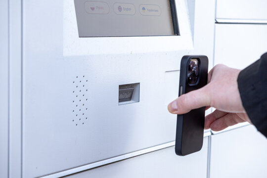 Man scanning smartphone at white parcel locker screen to collect package using QR code or app during modern contactless pickup process
