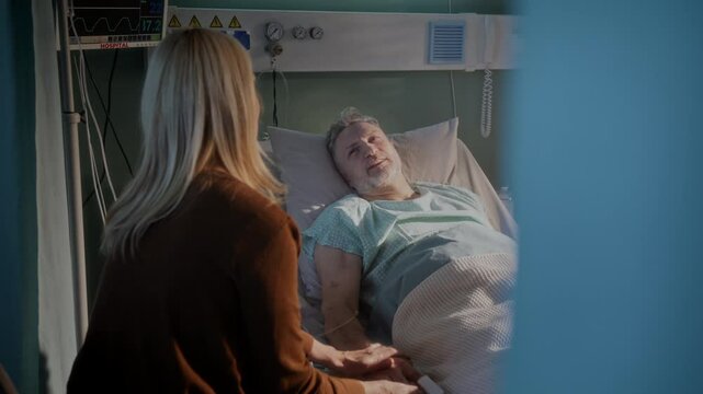 Woman sits beside bearded elderly man lying in hospital bed