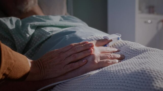 Wife strokes hand of elderly husband lying on hospital bed