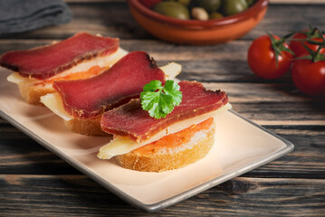 Slices of bread with tomato, topped with pieces of cured cheese and mojama, a typical tapa from Andalusia, salt-cured tuna loin following an ancestral tradition. Close-up.