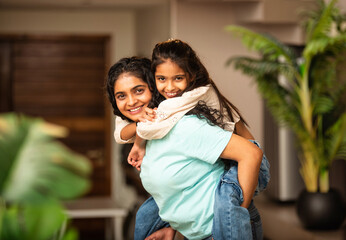 Cute Indian Asian girl enjoying a fun piggyback ride on her mother's back in a stylish living room
