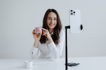 Young woman records personal finance tips while holding a piggy bank in a bright indoor setting,...