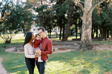 Smiling parents hugging little girl while standing with her in their arms in the park