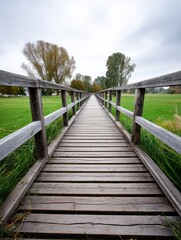 Obraz premium Wooden Bridge Path to Green Field - A long wooden bridge stretches towards a verdant field under a cloudy sky. Tranquil nature scene