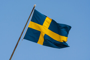 The blue and yellow flag of Sweden waving on a flagpole against a clear blue, sunny sky.