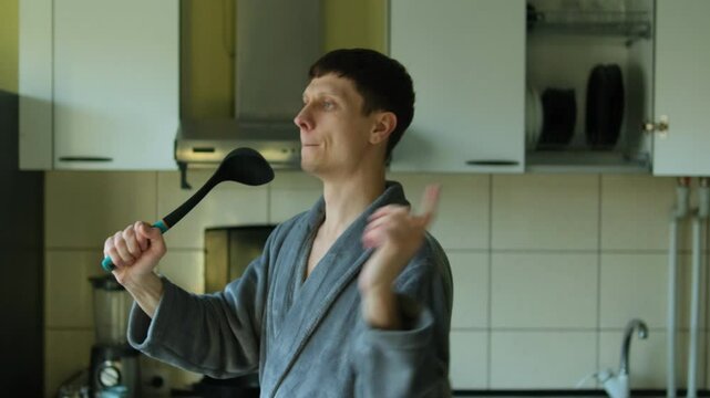 Man in a gray bathrobe playfully singing into a kitchen ladle like a microphone, expressing joy and lightheartedness in a modern kitchen environment enjoying culinary process. Slow motion