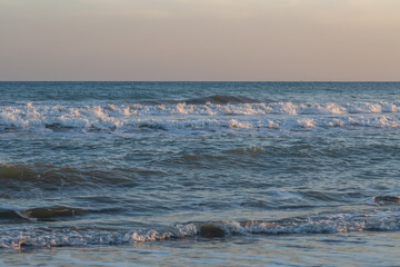 Obraz premium Crystal blue waves roll gently toward the shore under a pastel sky at sunset. The rhythmic movement of the sea creates a serene and calming atmosphere perfect for relaxation and reflection.