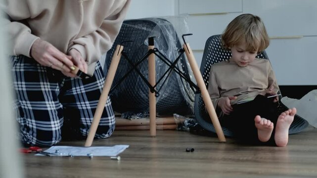 A man assembles a chair while his little son sits nearby and watches a tablet