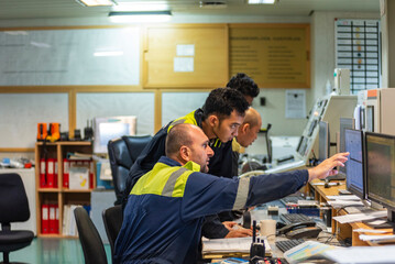 Marine engineering officers seated in the engine control room, focused on monitors in front of...