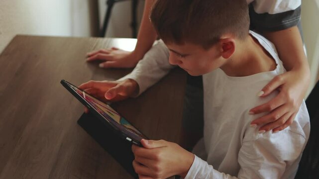 Supportive sister helping young brother play game or study on tablet, sharing warm moment of learning and connection at home with caring arm around his shoulder