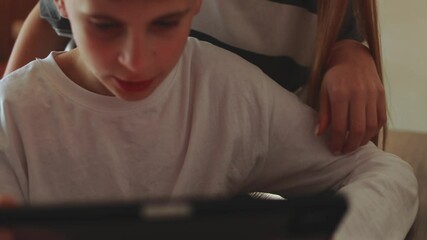 Supportive older sister resting hand on sibling's shoulder, watching younger brother concentrating deeply on digital tablet screen at home - Powered by Adobe