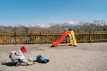 Little girl squatting digging sand with a toy shovel near a slide on a playground