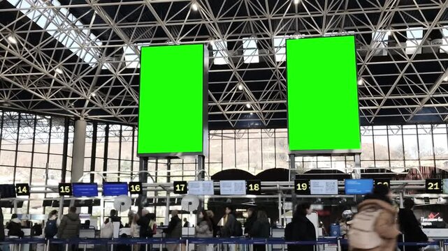Blank green screen billboard in a bustling airport terminal, with blurred passengers moving through the space.