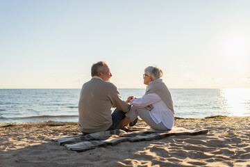 Senior mature couple sitting on beach holding hands enjoying outdoor recreation. Old husband wife touching hands with tenderness love. Grandmother grandfather together. Family moment of love