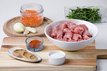Raw ingredients on a wooden cutting board against a white background pork meat, French mustard, white salt, hot pepper, onion, garlic, arugula, and red lentils. Fresh and natural food composition