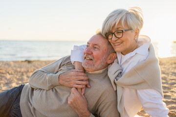 Senior mature couple sitting on beach hugging enjoying outdoor recreation. Old husband wife...