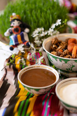 A beautifully arranged scene features bowls of nuts and dried fruits alongside a bowl of a brown liquid, possibly sauce. The items rest on a colorful, patterned textile with sprouts and little doll.