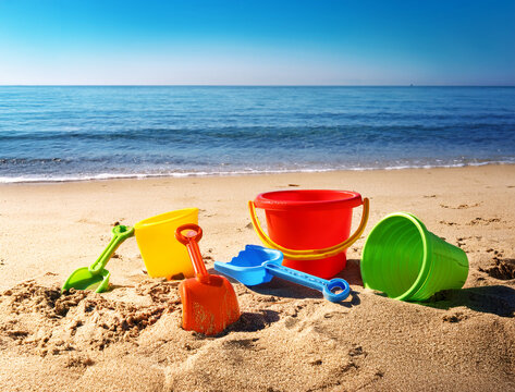 Colorful toy shovels, buckets, and sandcastles scattered on the beach sand with a calm sea and clear sky in the background, creating a peaceful, sunny atmosphere. - Powered by Adobe
