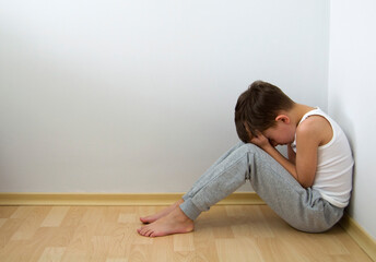 Boy crying with his head down and sitting sad scared on the floor in the corner and hugs his knees....