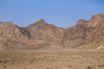 A high mountain range rises sharply from the plain. On the left is a wide strip of highway disappearing into the landscape. Shot in Yazd Province, Iran