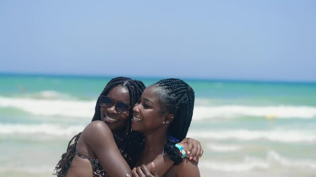 Two smiling friends share a joyful moment together hugging on a beach, enjoying the sunny atmosphere and cheerful vibes of the summer vacation, slow motion