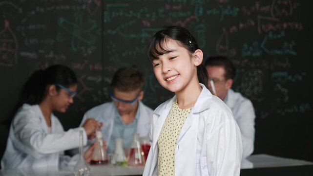 Asian scientist show miniheart to camera while diverse group doing experiment. Young schoolgirl standing at blackboard with chemical theory written while wearing lab coat at laboratory. Edification