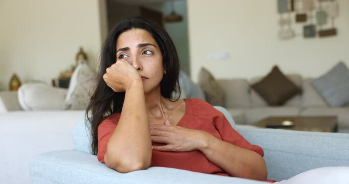 Depressed unhappy young Latin woman feeling bad, stressed, upset, touching head and chest, sitting in home armchair, suffering from headache, depression, apathy, thinking on loss, failure