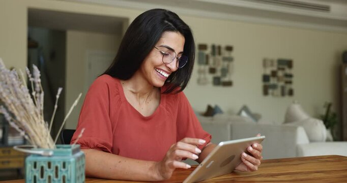Focused young Hispanic woman in stylish glasses using tablet computer at com table, watching online media content, getting happy, laughing, enjoying domestic Internet communication