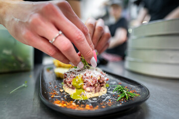 A chef's hands garnishing a finely chopped meat tartare with fresh herbs in a professional kitchen. The dish is elegantly presented on a black plate, complemented by sauce, grated cheese, and colorful