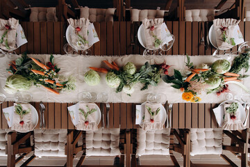Overhead view of a rustic wedding table setting with a wooden table, white runner, vegetable decor, and white plates