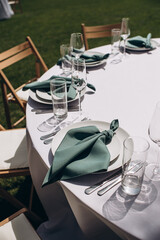 Close-up of an elegant outdoor wedding table setting with a white tablecloth and tied emerald green...