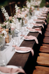 Elegant outdoor wedding table setting with a dark wooden table, white runner, pink napkins, and lush floral centerpieces