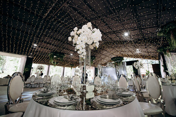 Elegant indoor wedding reception with round tables, mirrored tabletops, white tablecloths, tall floral centerpieces, and string lights