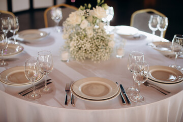 Elegant round wedding table setting with a white tablecloth, beige plates, simple white floral centerpiece, and tall wine glasses
