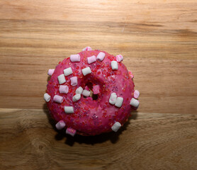 A bright pink donut garnished with white and pink marshmallows sits on a wooden cutting board. The warm kitchen ambiance enhances the appeal of this sweet treat