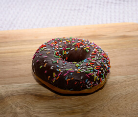 A round chocolate donut adorned with vibrant sprinkles rests on a wooden countertop, reflecting the warm ambiance of a cozy kitchen. Perfect for a sweet treat