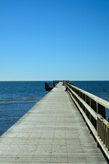 Obraz premium A wooden pier extends toward the horizon over a serene ocean. The clear blue sky creates a peaceful atmosphere, inviting visitors to enjoy the view.
