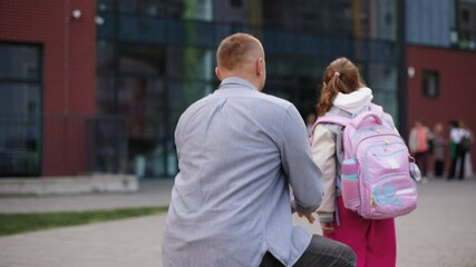 Father kneeling in front of his daughter, holding her by arms and looking at her with warmth and care. The girl is wearing backpack and a school jacket, standing in front of a modern school building.
