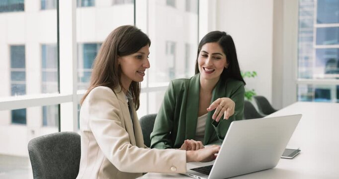 Two female business colleagues working at laptop together, talking at computer in office meeting room, discussing online project, Internet marketing strategy, pointing at pc screen, speaking