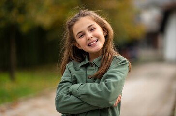 Portrait of a happy child girl with arms crossed outside, looking at the camera and smiling.