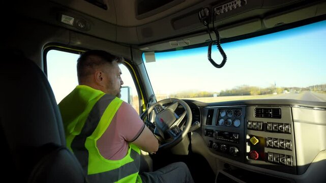 Young handsome Professional Semi Truck Driver wearing safety vest is riding a truck on the beautiful sunny day. Shot from inside the truck