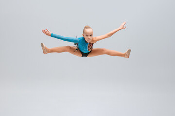 A young gymnast in a blue and leopard-print leotard performs a powerful split jump in mid-air. Her arms are extended, expression focused, and the clean background highlights her athleticism.