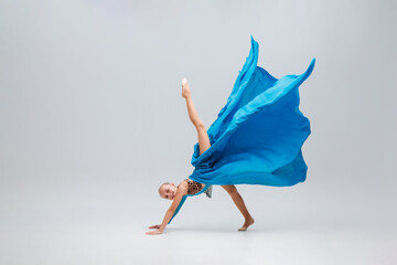 ballerina performing an acrobatic move stands on her hands with a leg raised high. Cheerleader is wearing a sport leotard and a flowing blue cloth, light gray background accentuates dynamic motion.