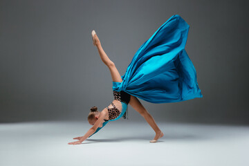 Cheerleader dancing with flying blue cloth fabric. Graceful ballerina dancing on grey studio background. Dancer in sport leotard doing Split. Child, artist, movement, action and motion concept